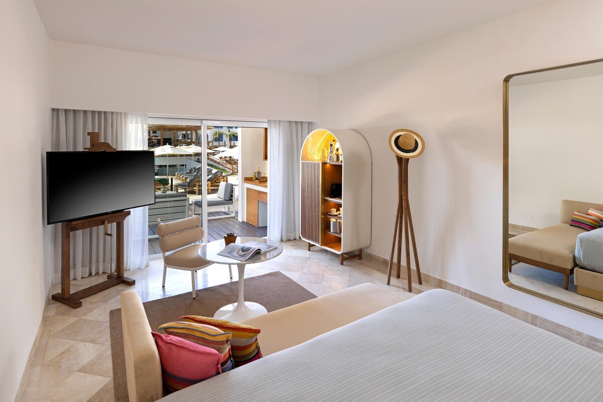 A hotel room with a TV, a bed, a sofa, a dining table, a mirror, and a view of the outdoor area.