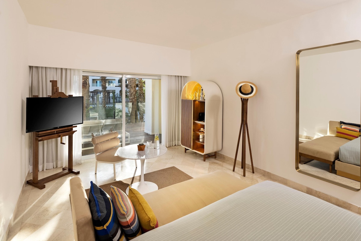 A hotel room with a bed, a TV, a seating area with a table, a mirror, and a view of the outdoors.
