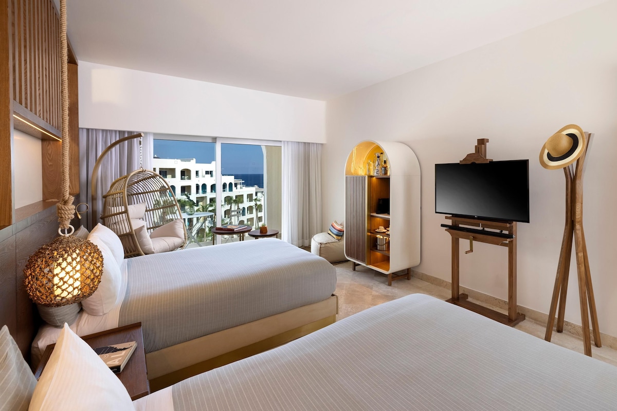 A hotel room with two beds, a TV, a wardrobe, a hanging chair, and a view of buildings.