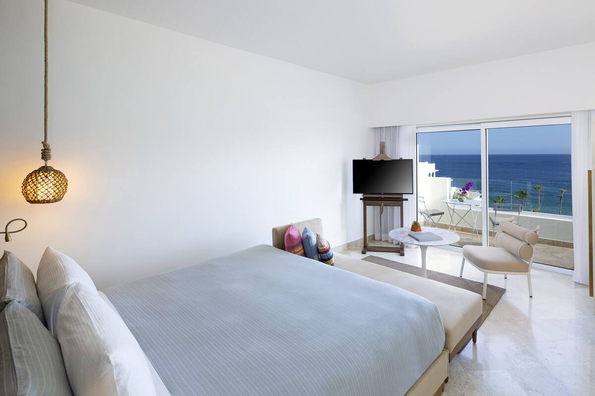 Habitación de hotel con cama, televisor, balcón con vistas al mar y un sombrero en un perchero.