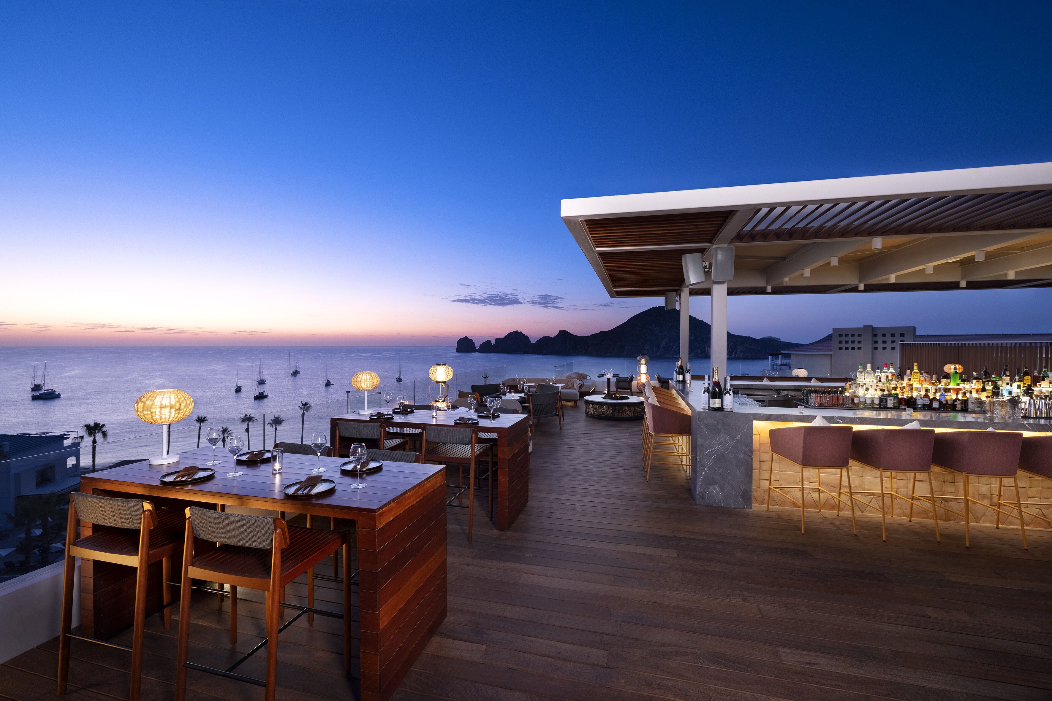 Un bar en la azotea con mesas y sillas de madera, zona de bar con taburetes y vistas al océano y a los barcos al atardecer.
