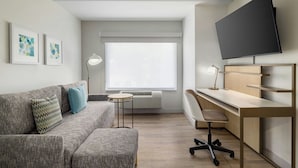 Una habitación moderna y bien iluminada con un sofá gris, una pequeña mesa redonda, un escritorio con una silla, un televisor montado y obras de arte en la pared.