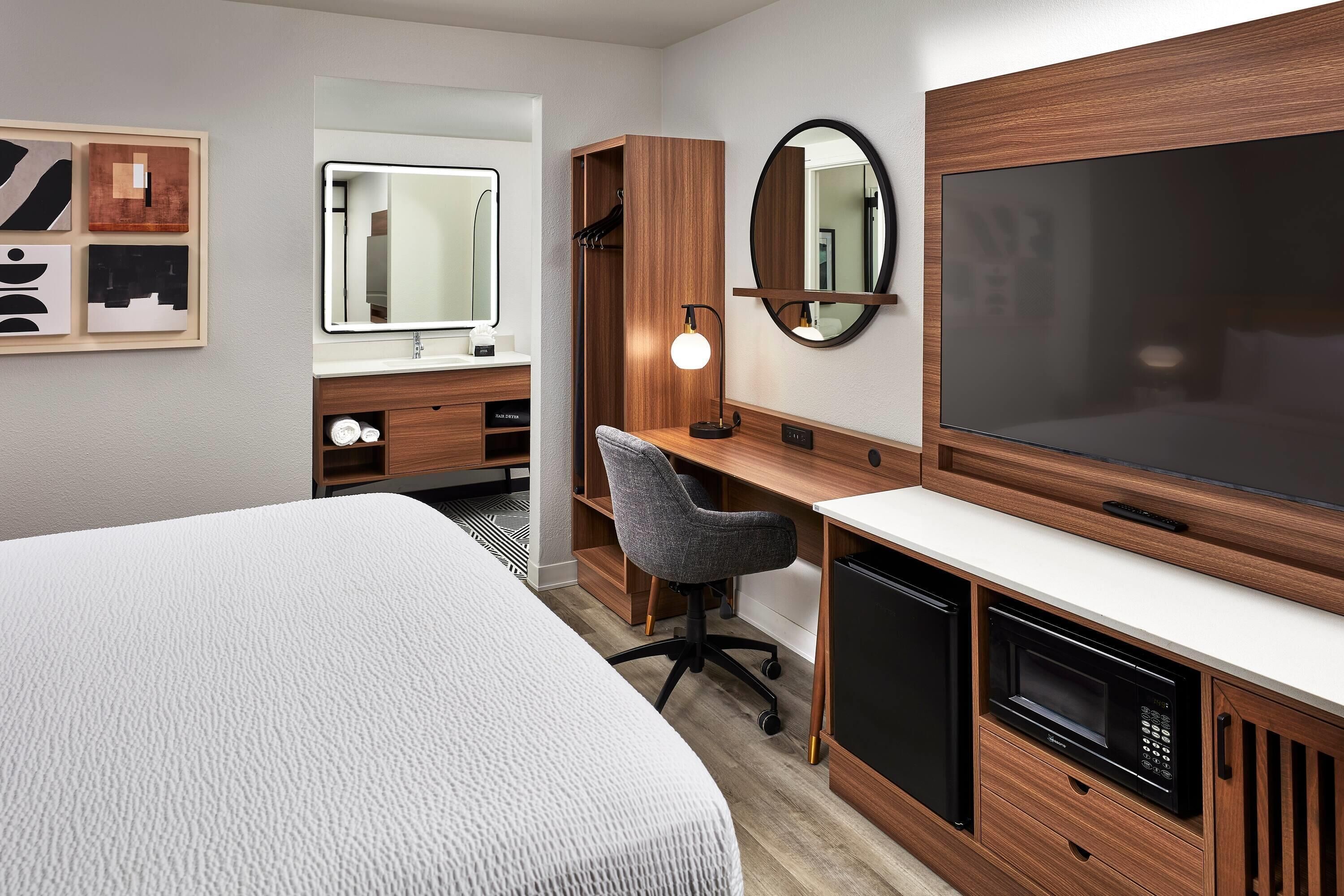 A modern hotel room with a large bed, a desk with a chair, a TV, a mirror, and a wardrobe.