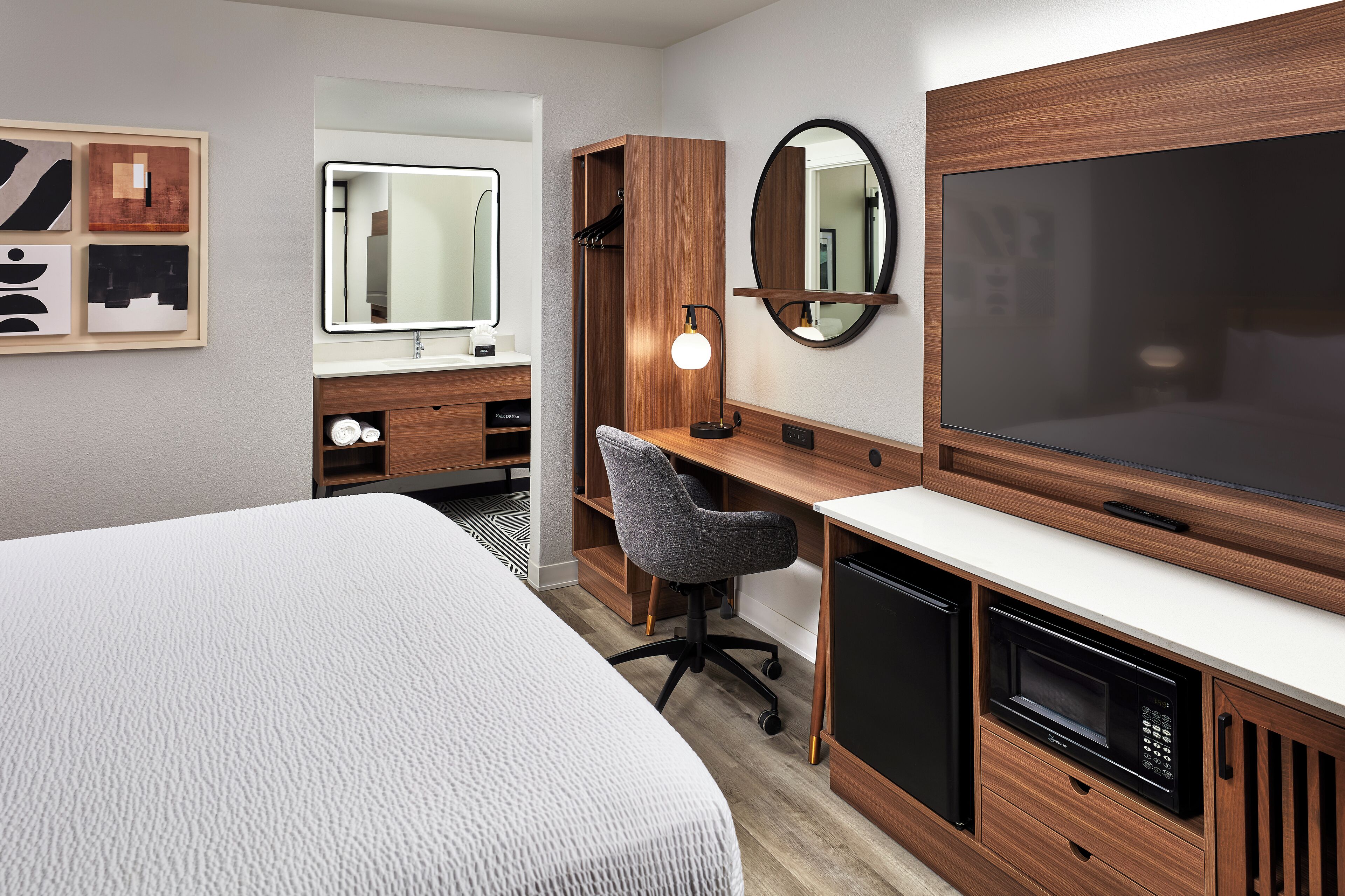 A modern hotel room with a large bed, a desk with a chair, a television, a mirror, and a wardrobe.