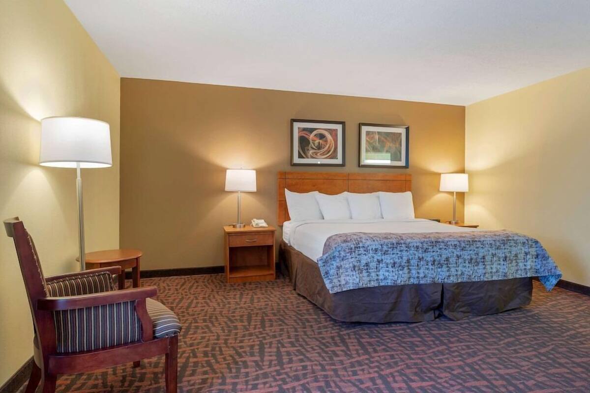 A hotel room with a large bed, two bedside lamps, a wooden nightstand, a chair, and two framed pictures on the wall.