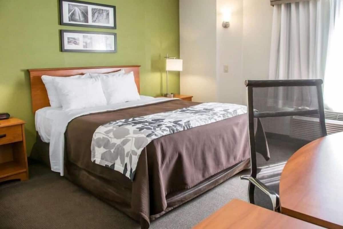 A hotel room with a bed, a chair, a table, a lamp, and two framed pictures on the wall.