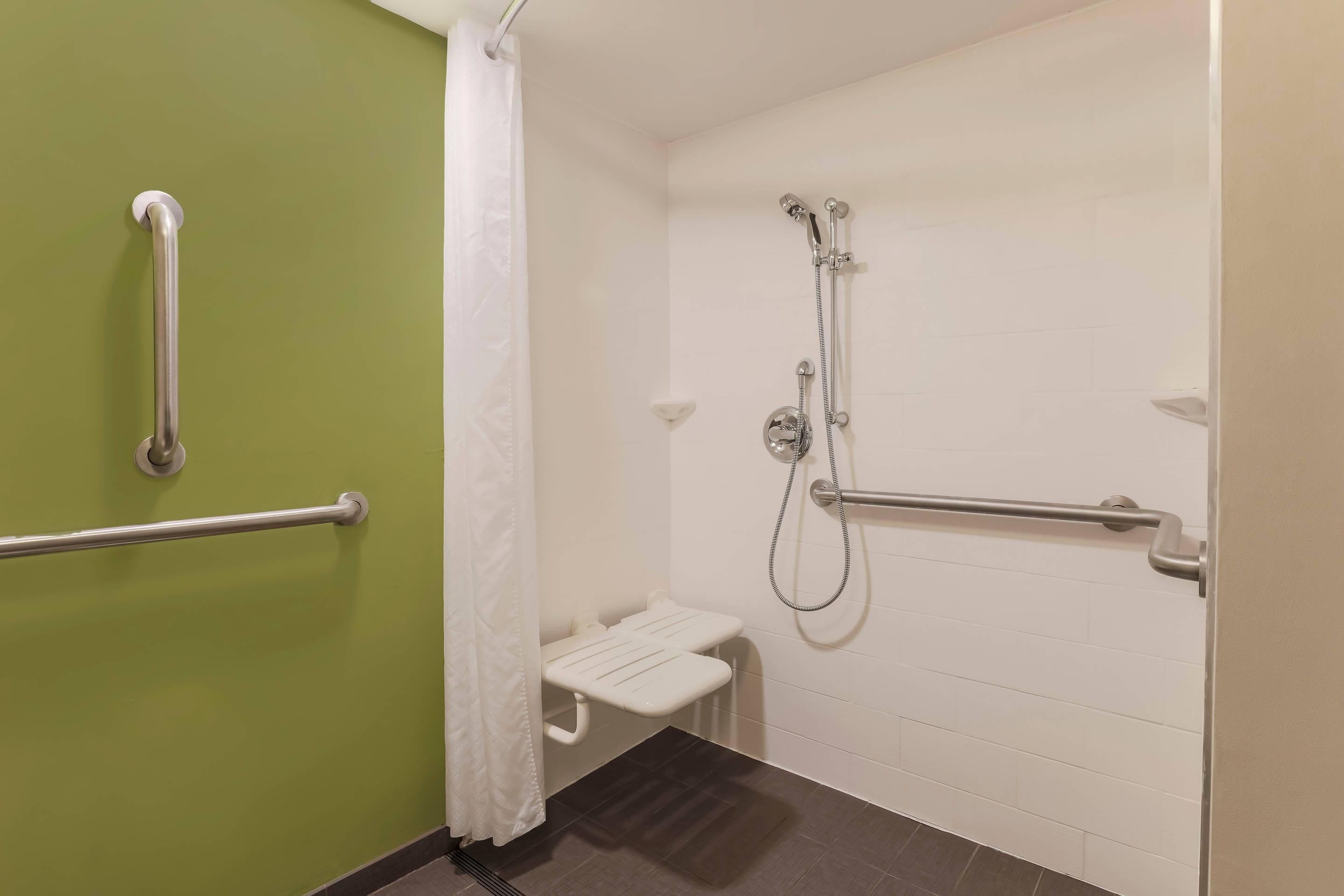 A bathroom with a shower, grab bars, a white bench, and a green wall.