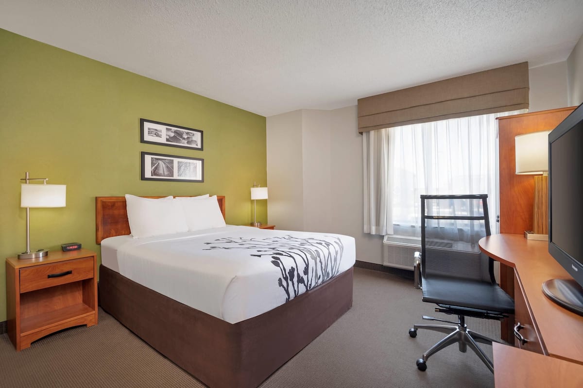 A hotel room with a large bed, bedside tables with lamps, a desk with a chair, a television, and framed pictures on the wall.