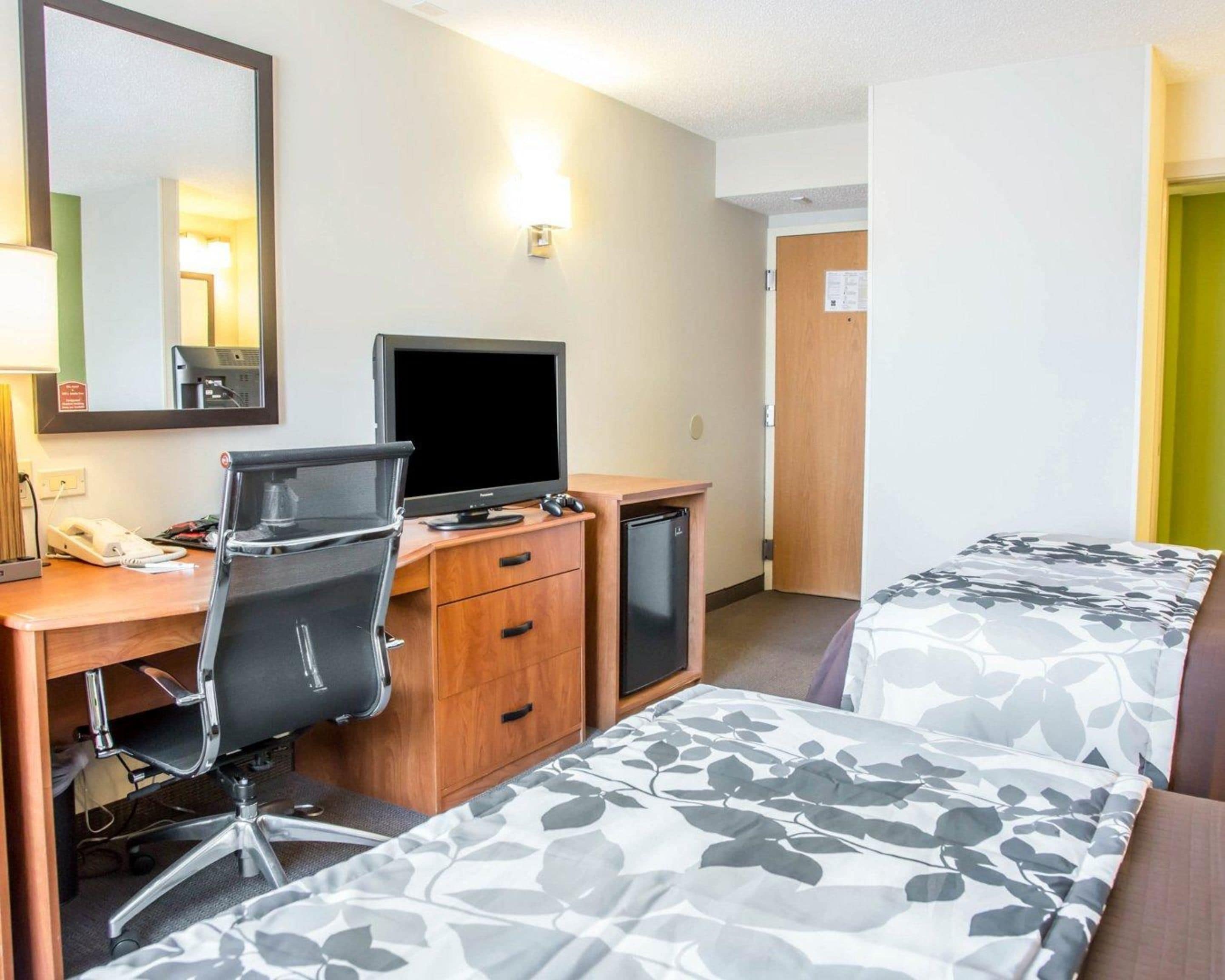 A hotel room with a desk, a chair, a bed with floral bedding, a television, and a small refrigerator.