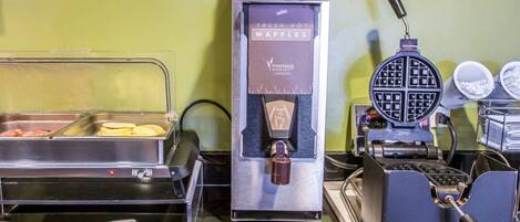 A buffet setup with a waffle maker, a hot food dispenser, and a tray of food items.
