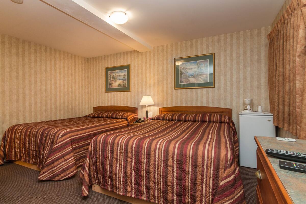 Une chambre d’hôtel avec deux lits, un petit bureau, une lampe, un réfrigérateur et deux tableaux encadrés au mur.