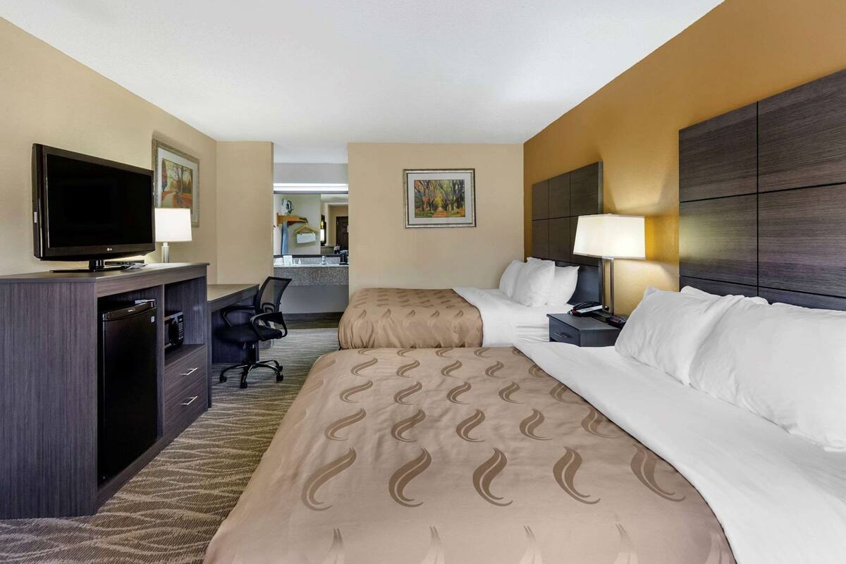 A hotel room with two beds, a desk with a TV, a chair, and a bathroom visible in the background.