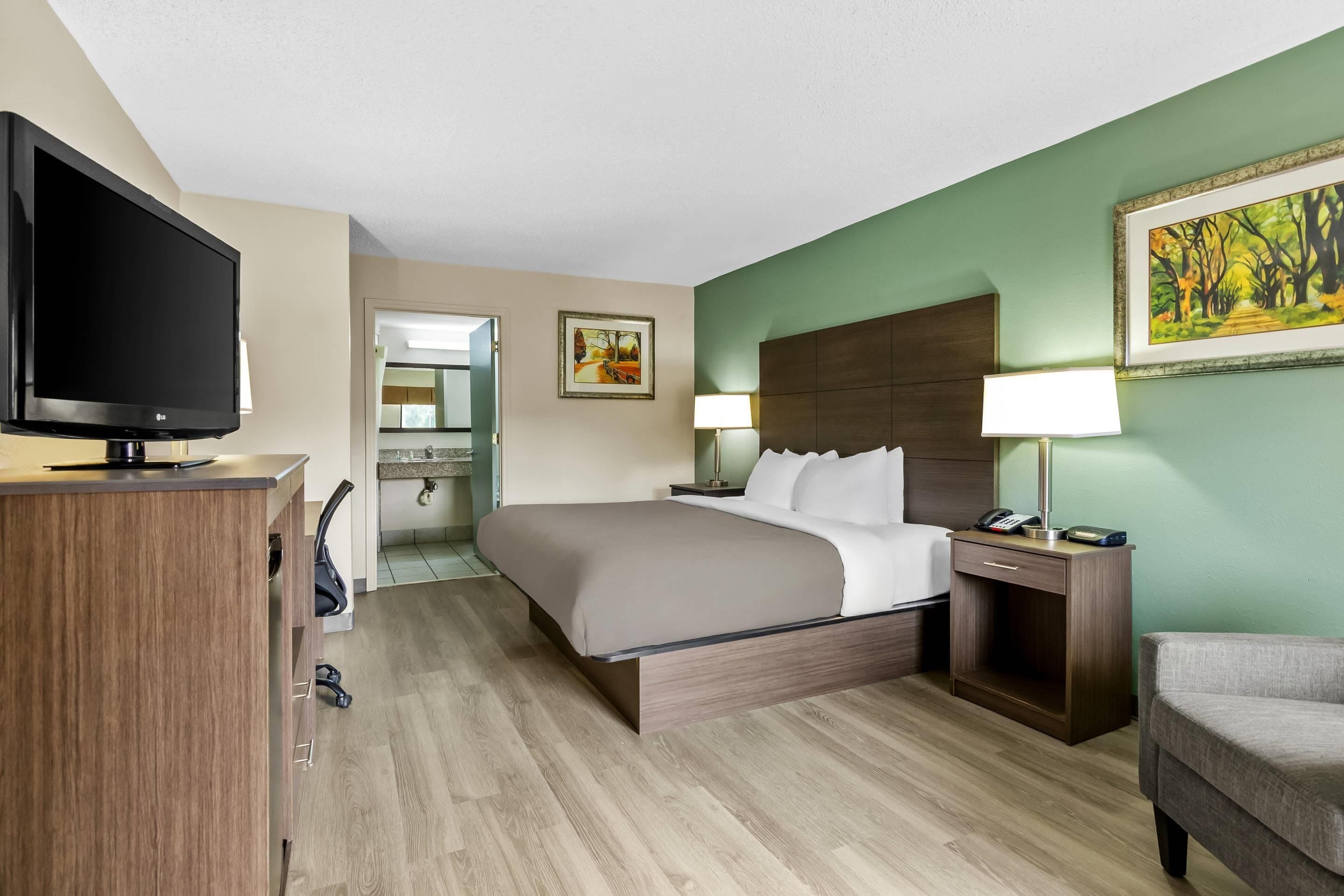 Habitación de hotel con cama, televisor, escritorio, silla, mesita de noche y un cuadro en la pared.