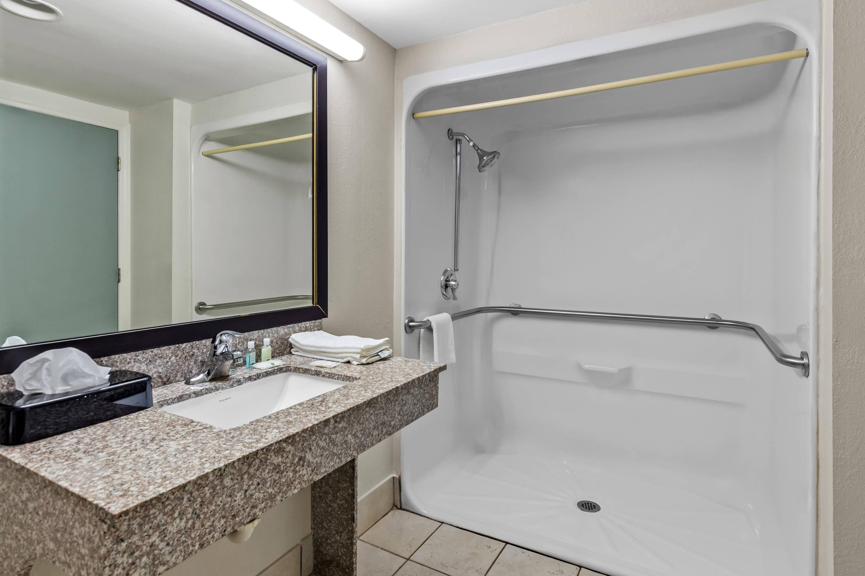 Un baño con encimera de granito, un lavabo, un espejo, ducha con barra de apoyo y un perchero para toallas.