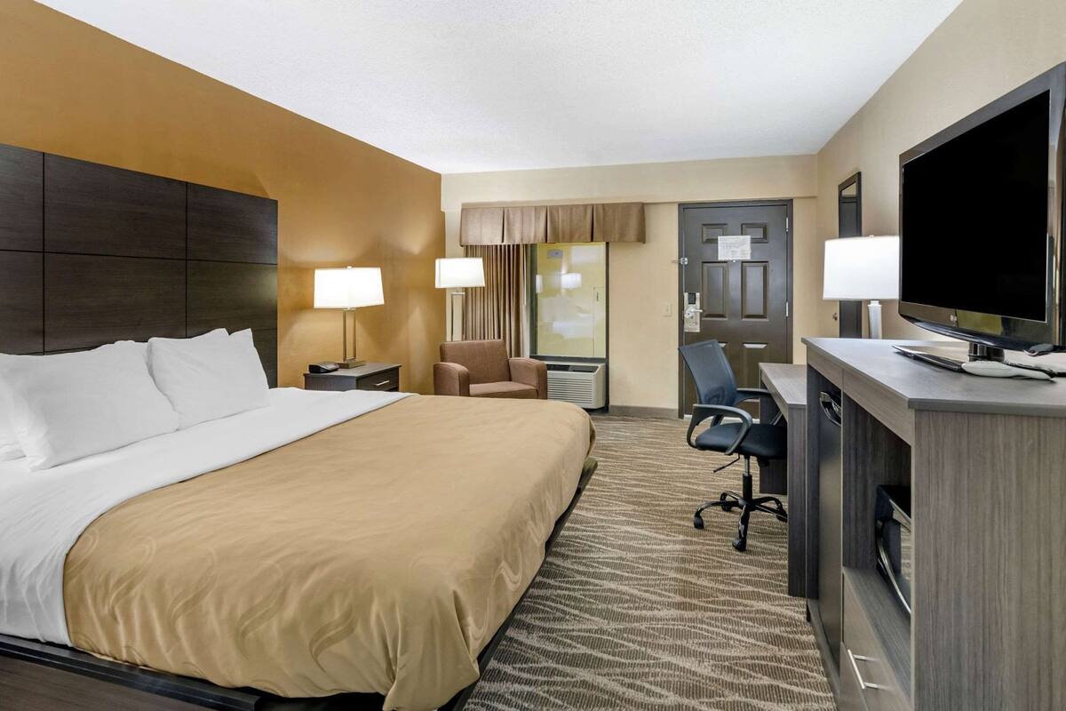 A hotel room with a bed, two chairs, a desk with a TV, and a minibar.
