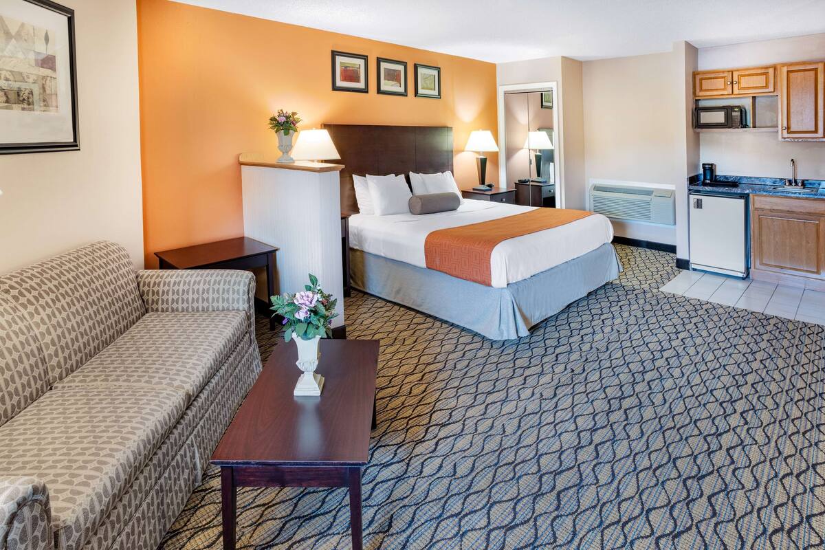 A hotel room with a bed, a sofa, a small table with a vase, a kitchenette, and a patterned carpet.