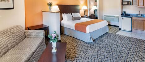 Ein Hotelzimmer mit Bett, Sofa, einem kleinen Tisch mit Vase, einer Küchenzeile und einem gemusterten Teppich.