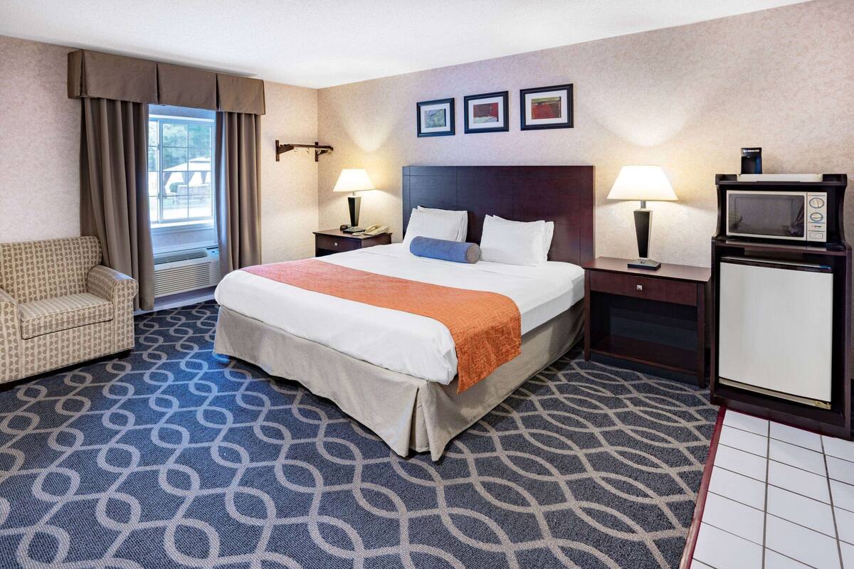 A hotel room with a bed, a chair, a desk with a lamp, a microwave, and a small refrigerator.