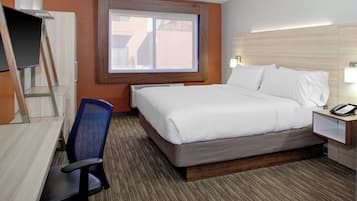 A hotel room with a bed, a desk, a chair, a window, and a bedside table with a lamp.