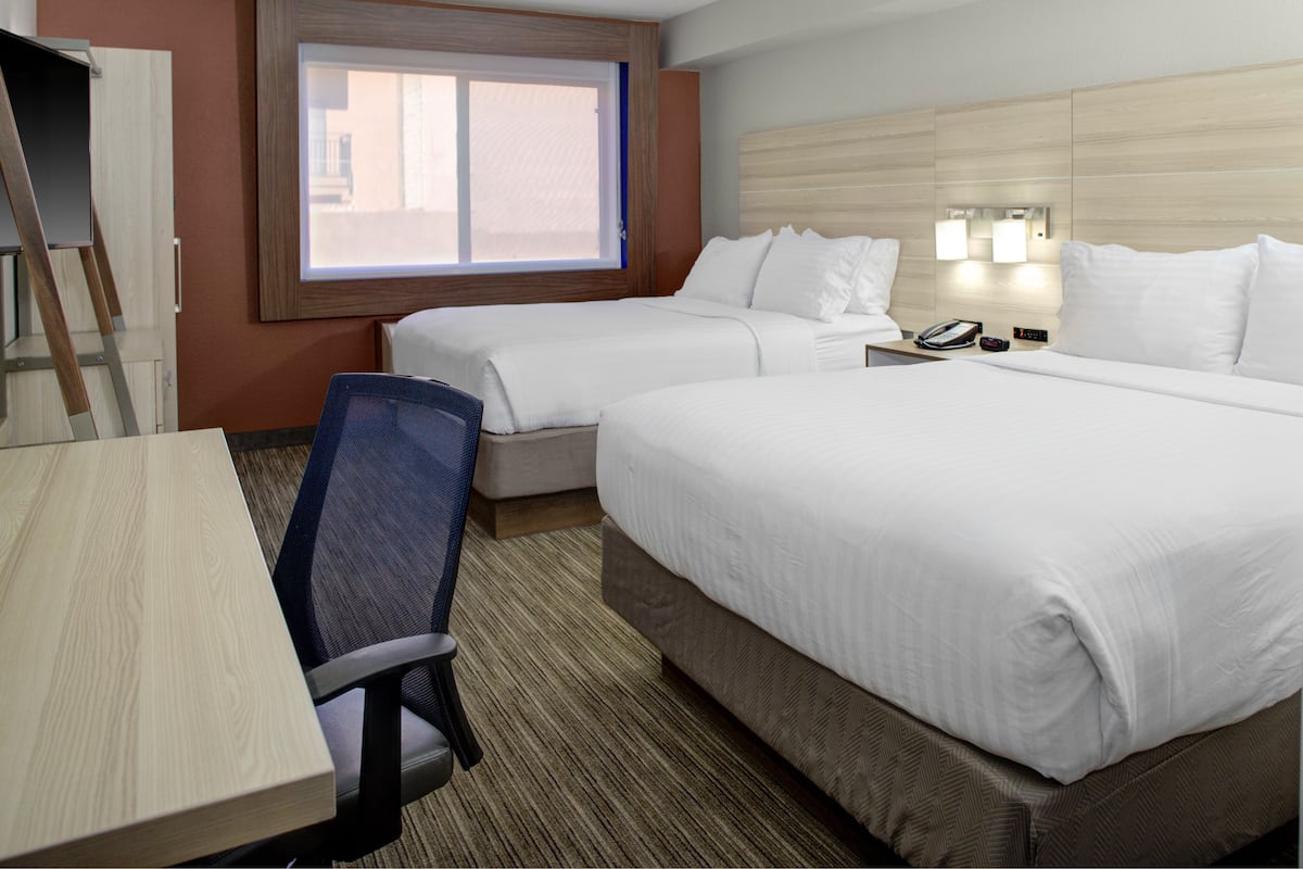 A hotel room with two beds, a desk, a chair, a window, and a nightstand with a phone and alarm clock.