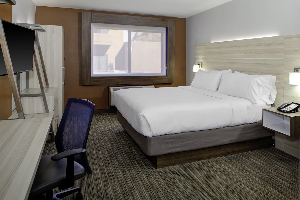 A hotel room with a bed, a desk, a chair, a window, and a nightstand with a phone.