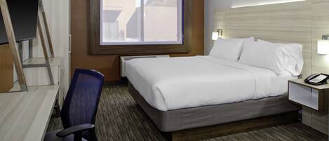 A hotel room with a bed, a desk, a chair, a window, and a nightstand with a phone.