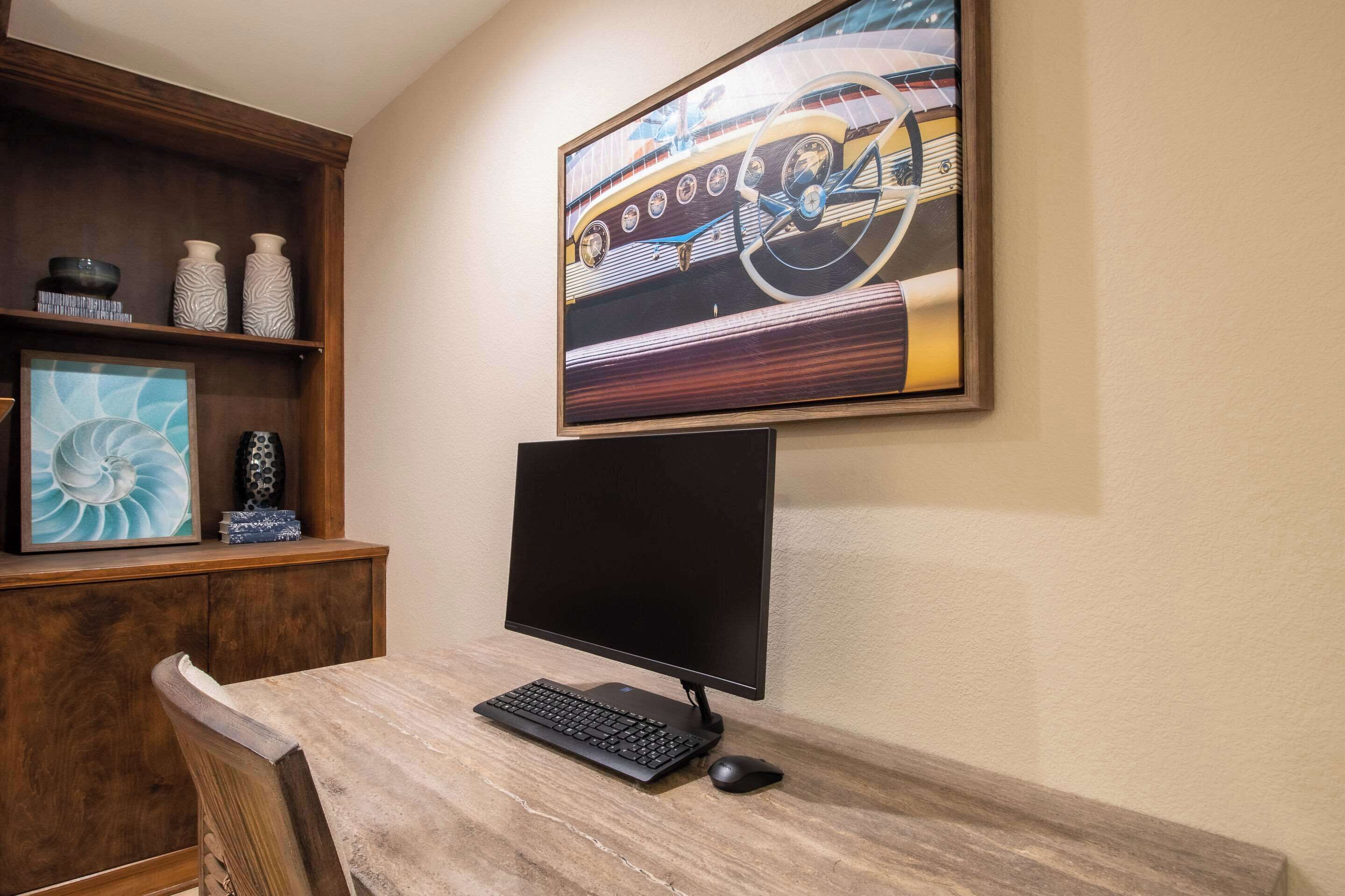 Un escritorio de madera con una computadora, que incluye un monitor, teclado y ratón. Sobre el escritorio, hay una obra de arte enmarcada de un coche antiguo.