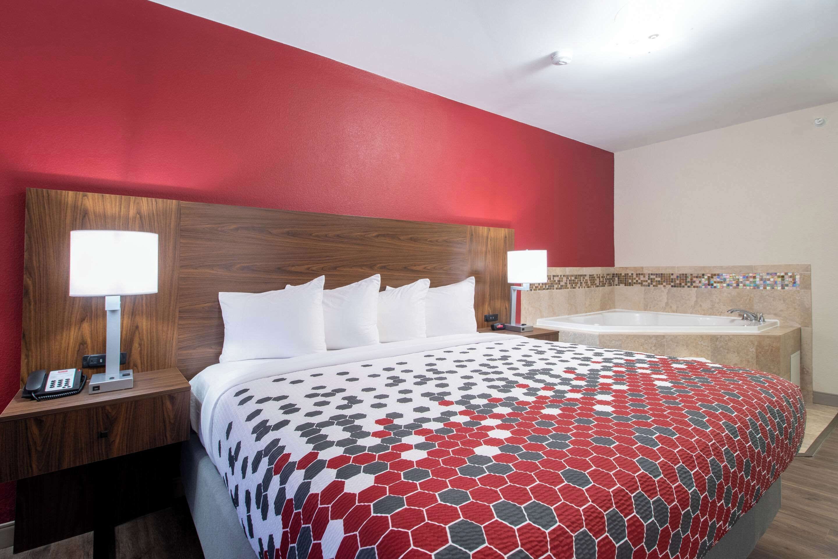 Habitación de hotel con una cama grande, dos lámparas de noche, un cabecero de madera, una pared de acento roja, un baño con bañera y zona de ducha con azulejos.