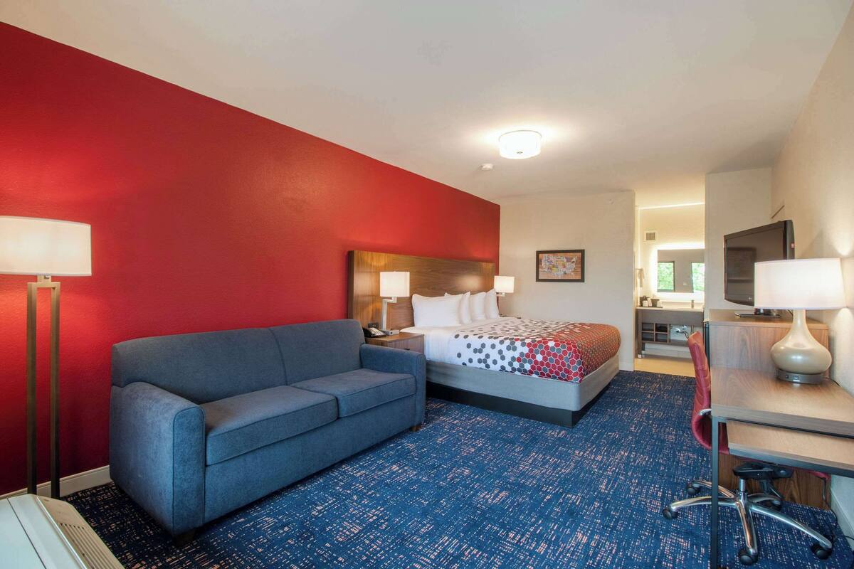Een hotelkamer met een rode accentmuur, een blauw kamerkleed met patroon, een bed met een stippenkleed, een grijze bank, een bureau met stoel en een televisie aan de muur.