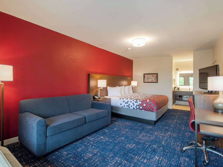A hotel room with a red accent wall, a blue patterned carpet, a bed with a polka dot blanket, a gray sofa, a desk with a chair, and a TV on the wall.