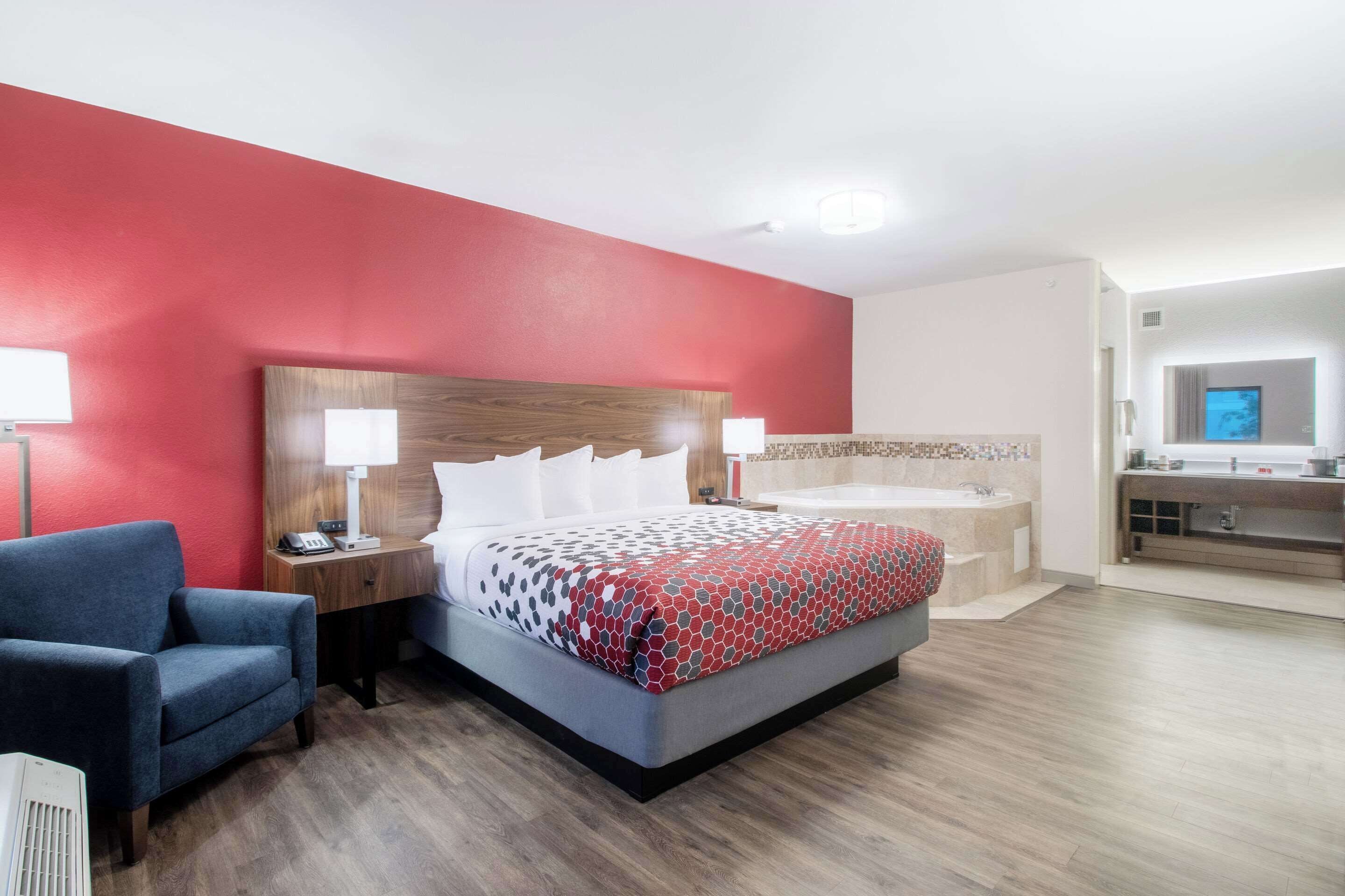A hotel room with a bed, a chair, a desk, a lamp, a bathroom with a bathtub, and a mirror.