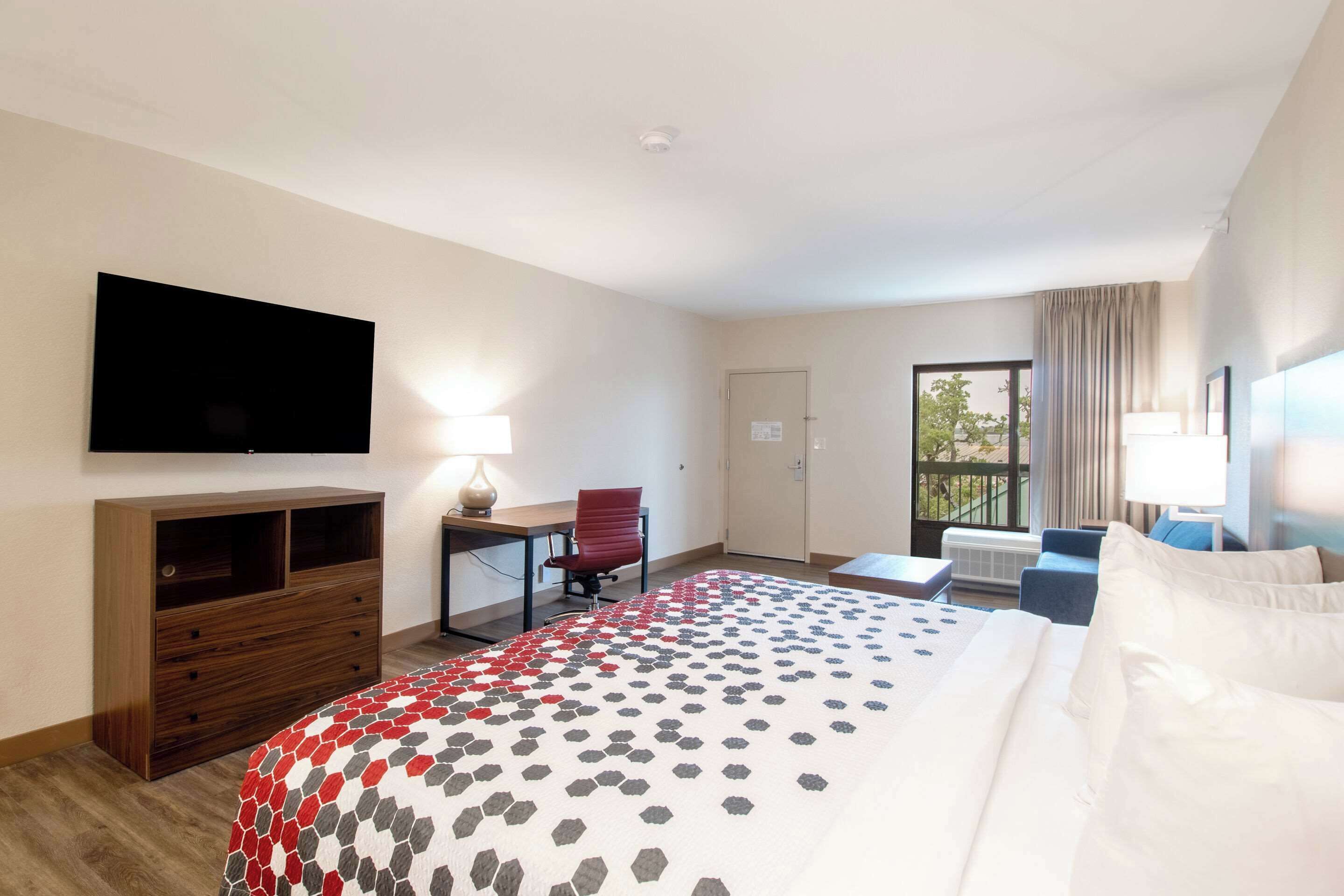A hotel room with a large bed, a desk with a chair, a flat-screen TV mounted on the wall, and a window with curtains.