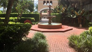 A courtyard with a central fountain, surrounded by lush greenery and a building with arched windows in the background. - Grand Palms Spa & Golf Resort (Pembroke Pines)