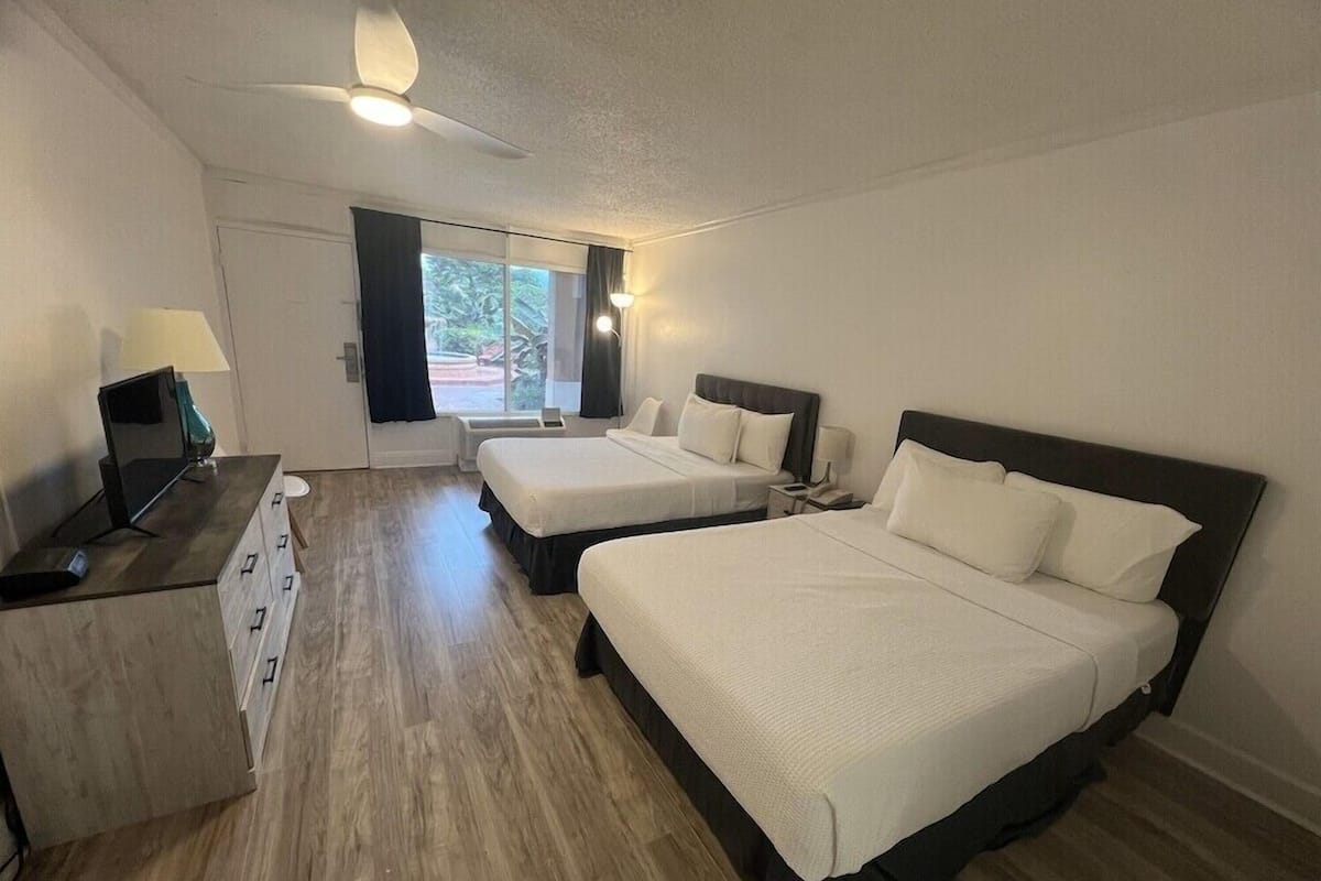 A hotel room with two beds, a desk with a computer, a ceiling fan, and a window with curtains.