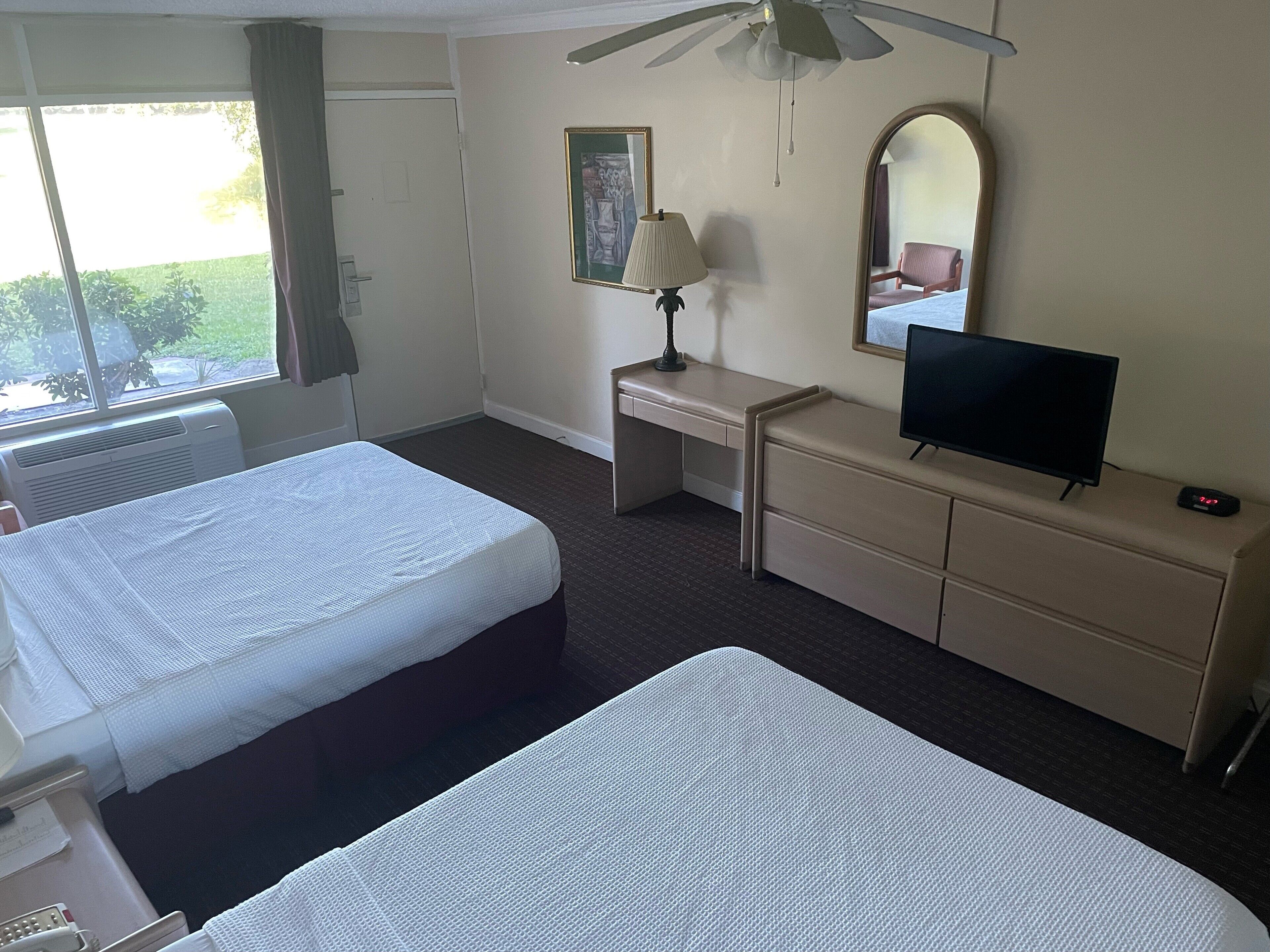Una habitación de hotel con dos camas, un escritorio, televisor, espejo y ventana con cortinas.