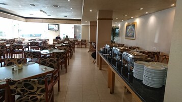 Un amplio comedor con varias mesas y sillas, un mostrador de buffet con platos apilados y un televisor montado en la pared.