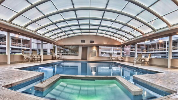 An indoor swimming pool with a glass ceiling, surrounded by lounge chairs and a tiled floor.
