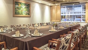 A conference room with a long table set for a meeting, wooden chairs, a large window with a view of parked cars, and a framed painting on the wall.