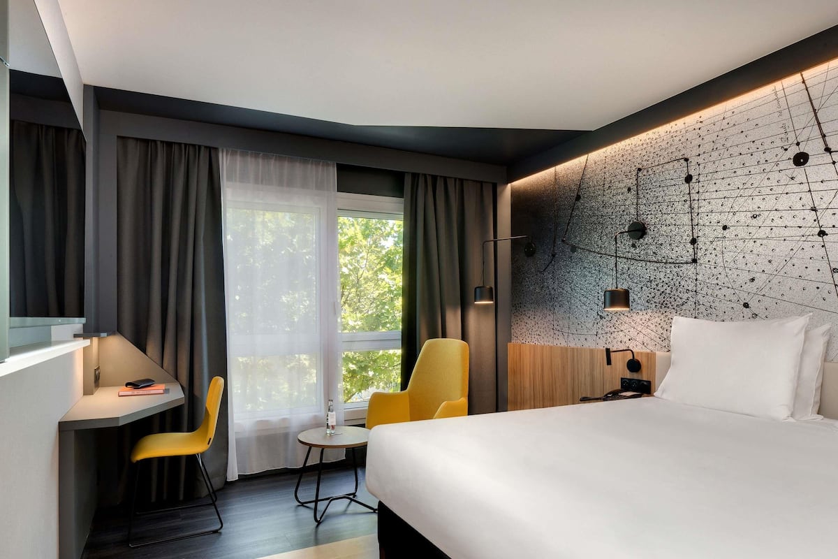 A modern hotel room with a large bed, a desk, a chair, and a view of trees through the window.