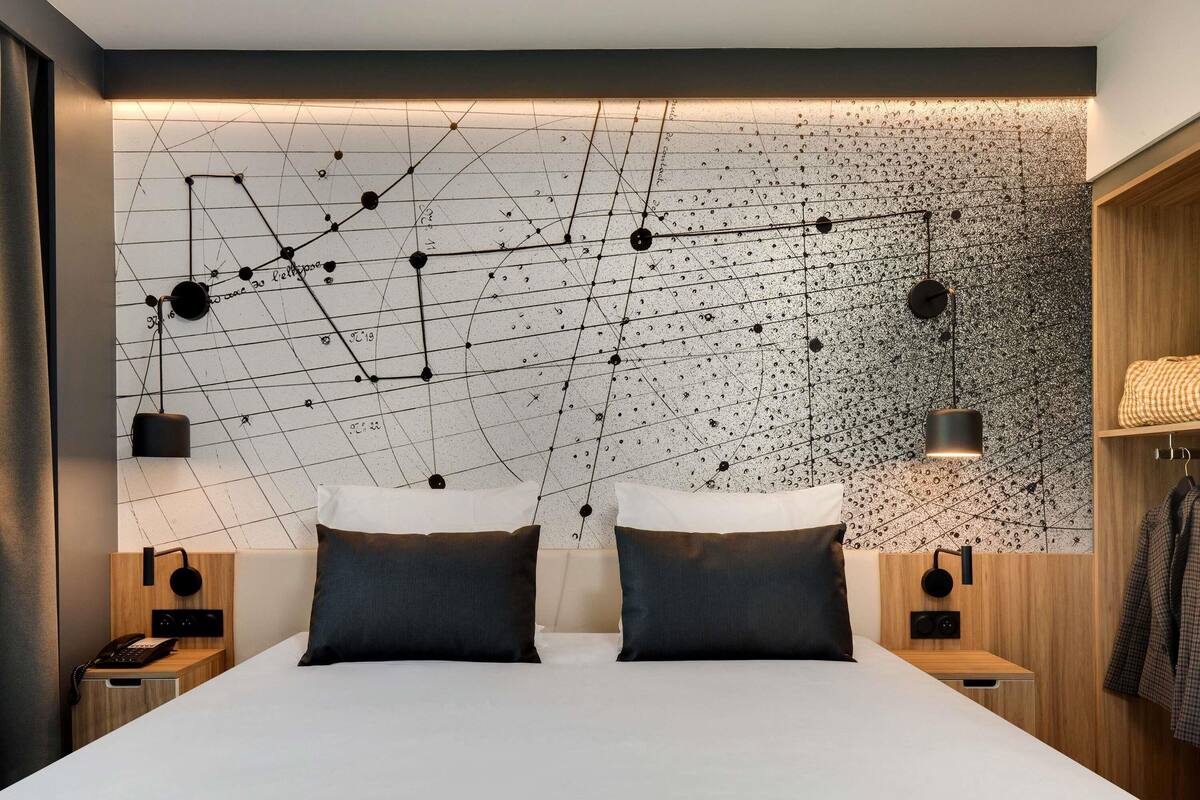 A modern bedroom with a geometric wall design, two bedside tables with lamps, a bed with two pillows, and a wooden wardrobe.