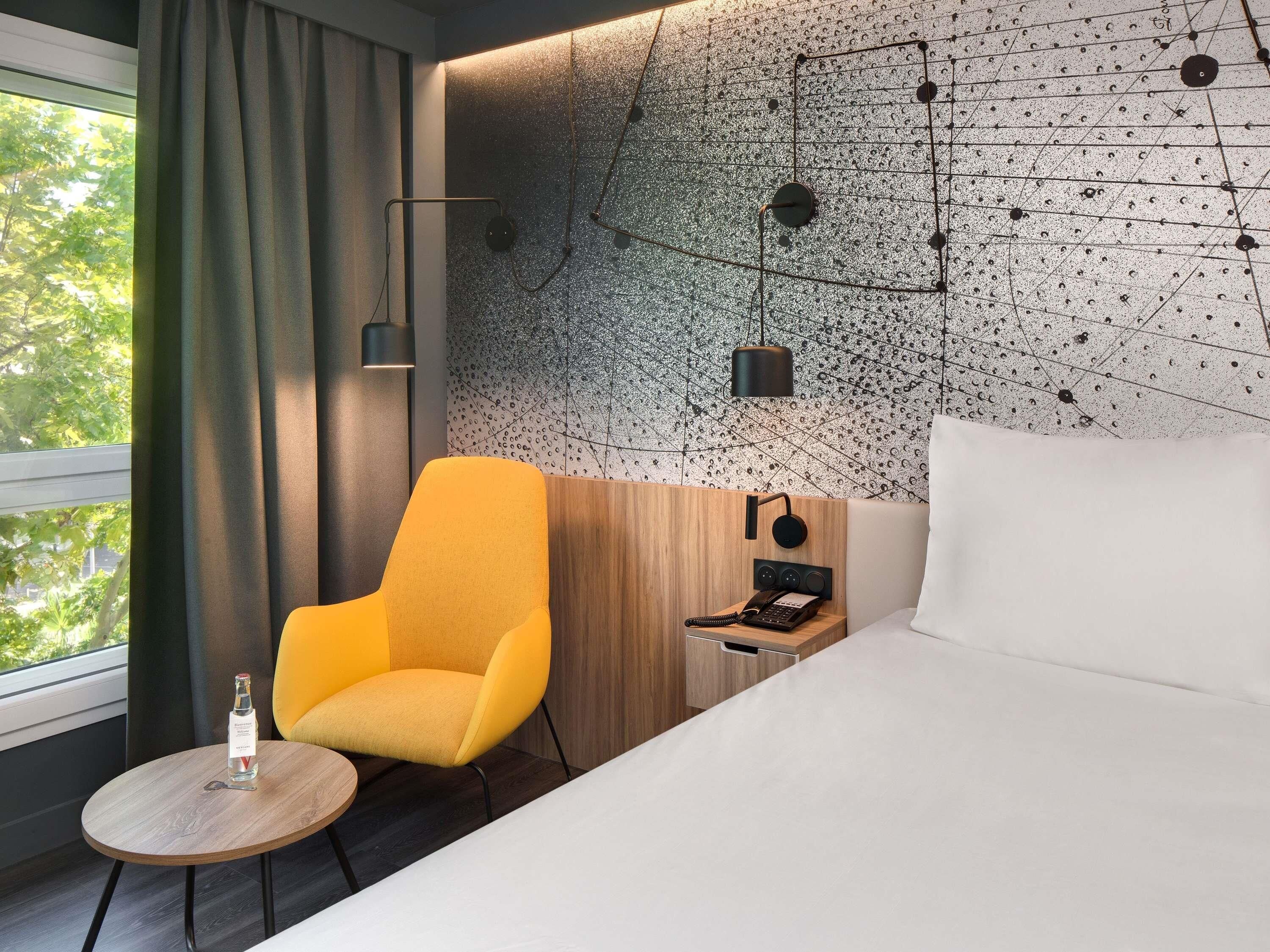 A hotel room with a bed, a yellow armchair, a small table with a bottle, a wall-mounted phone, and a decorative wall with a geometric pattern.