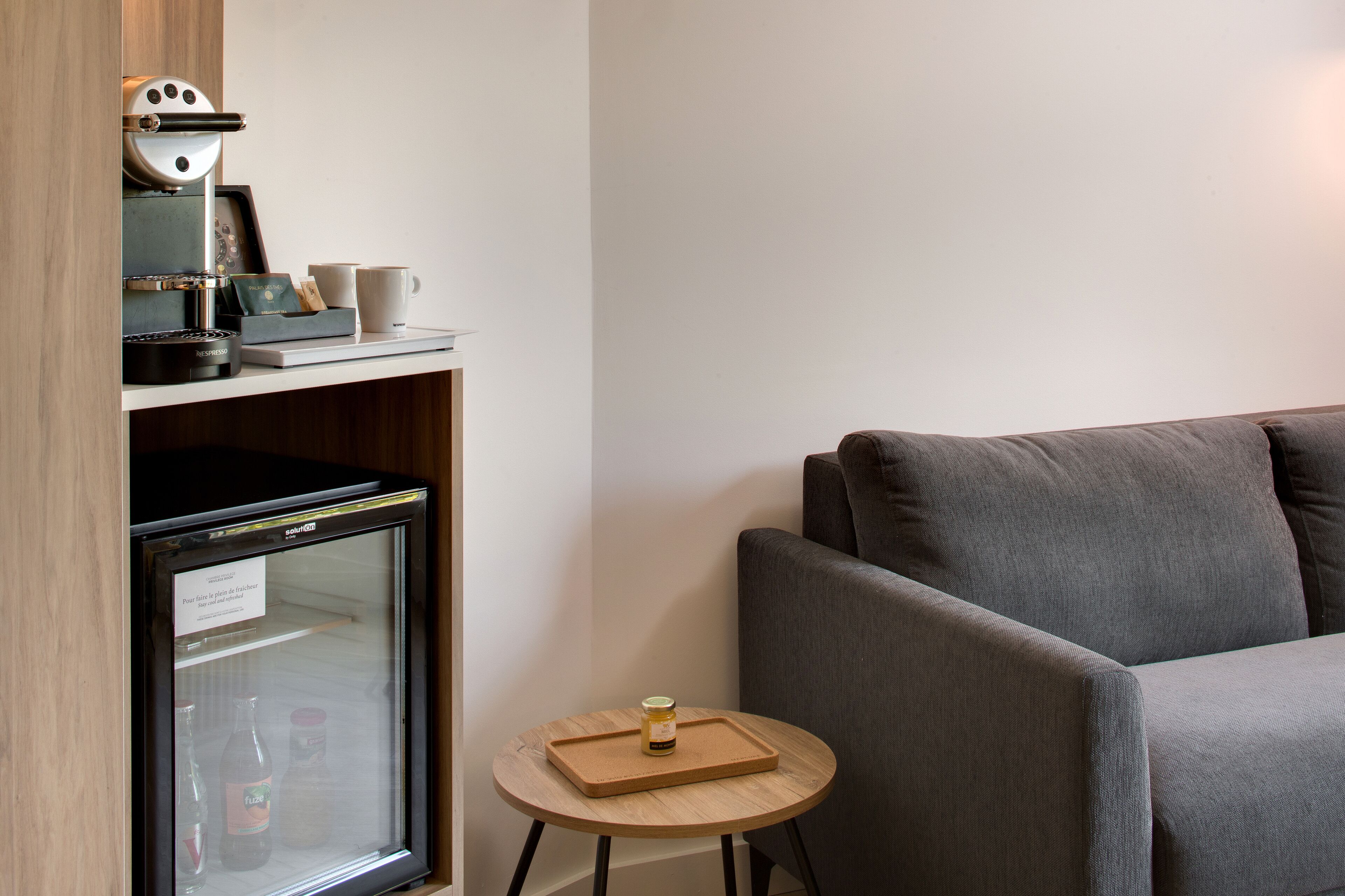 Une pièce de taille modeste, équipée d’un réfrigérateur, d’une petite table avec un plateau et d’un canapé gris.