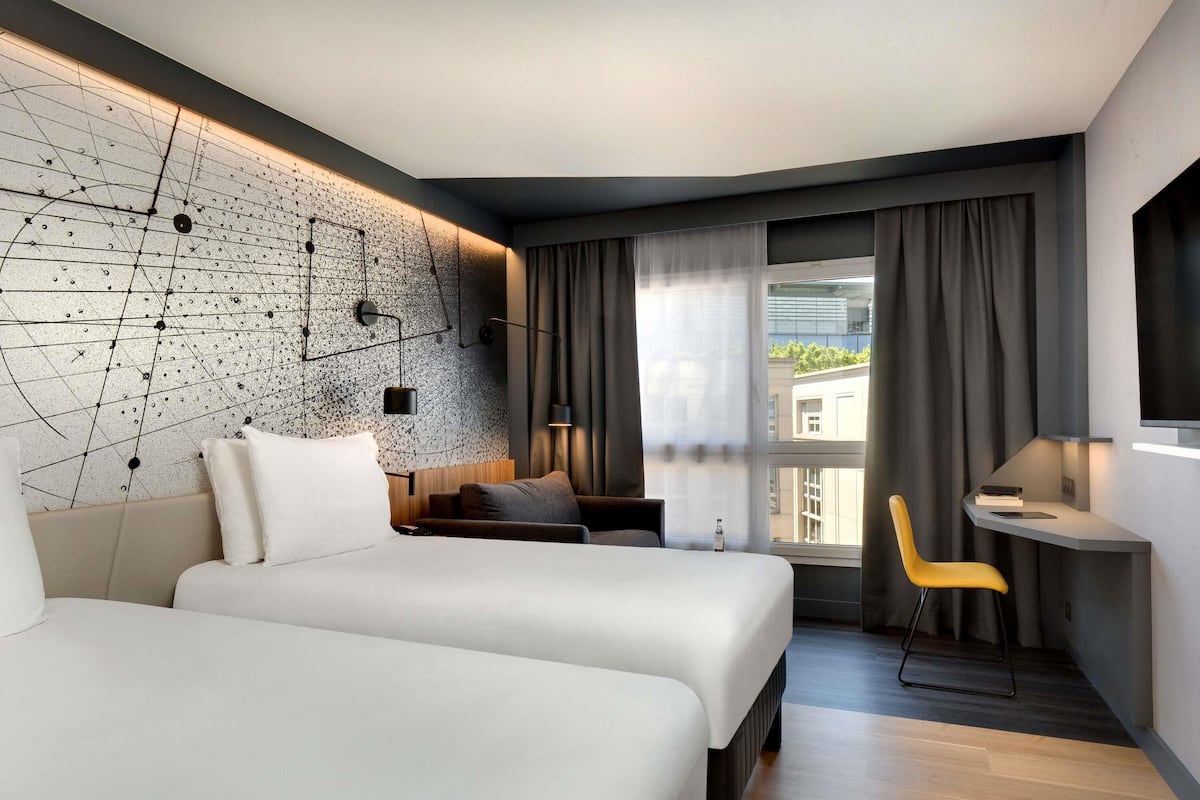 A modern hotel room with two beds, a desk, a chair, a large window, and a unique wall design.