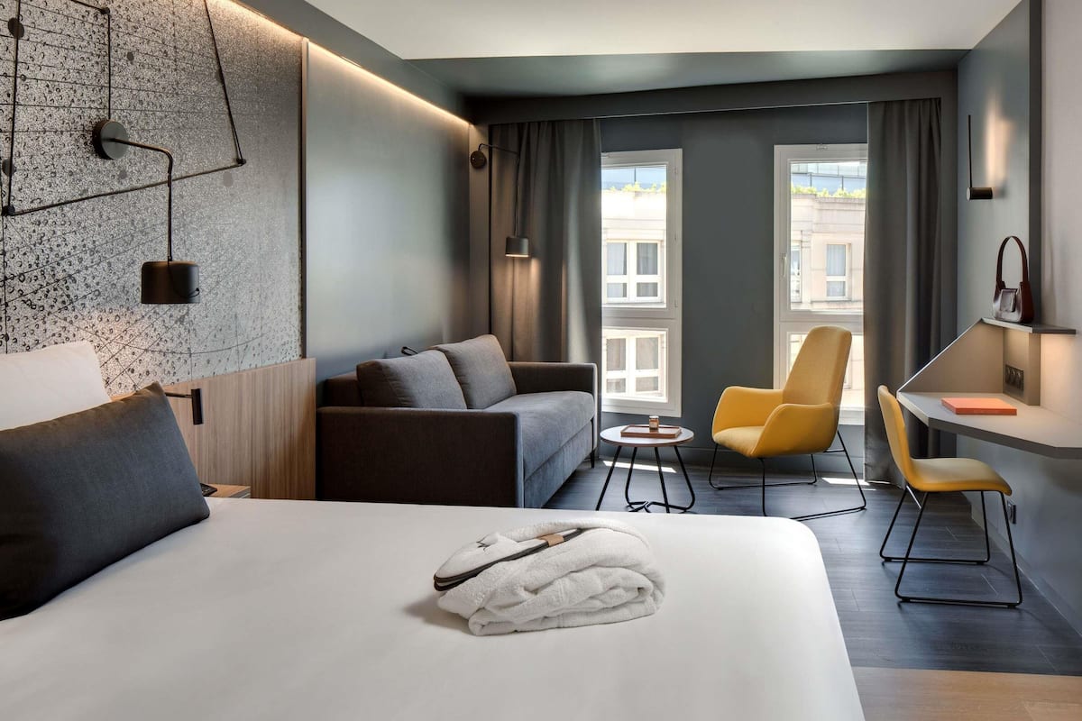 A modern hotel room with a large bed, a sofa, a chair, a small table, and a desk.