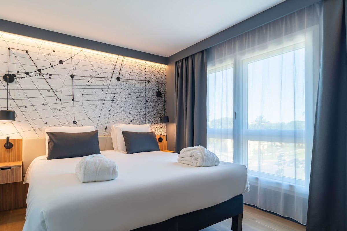 Un hotel moderno con una cama grande, un diseño geométrico en la pared y vistas de la ciudad a través de amplias ventanas.