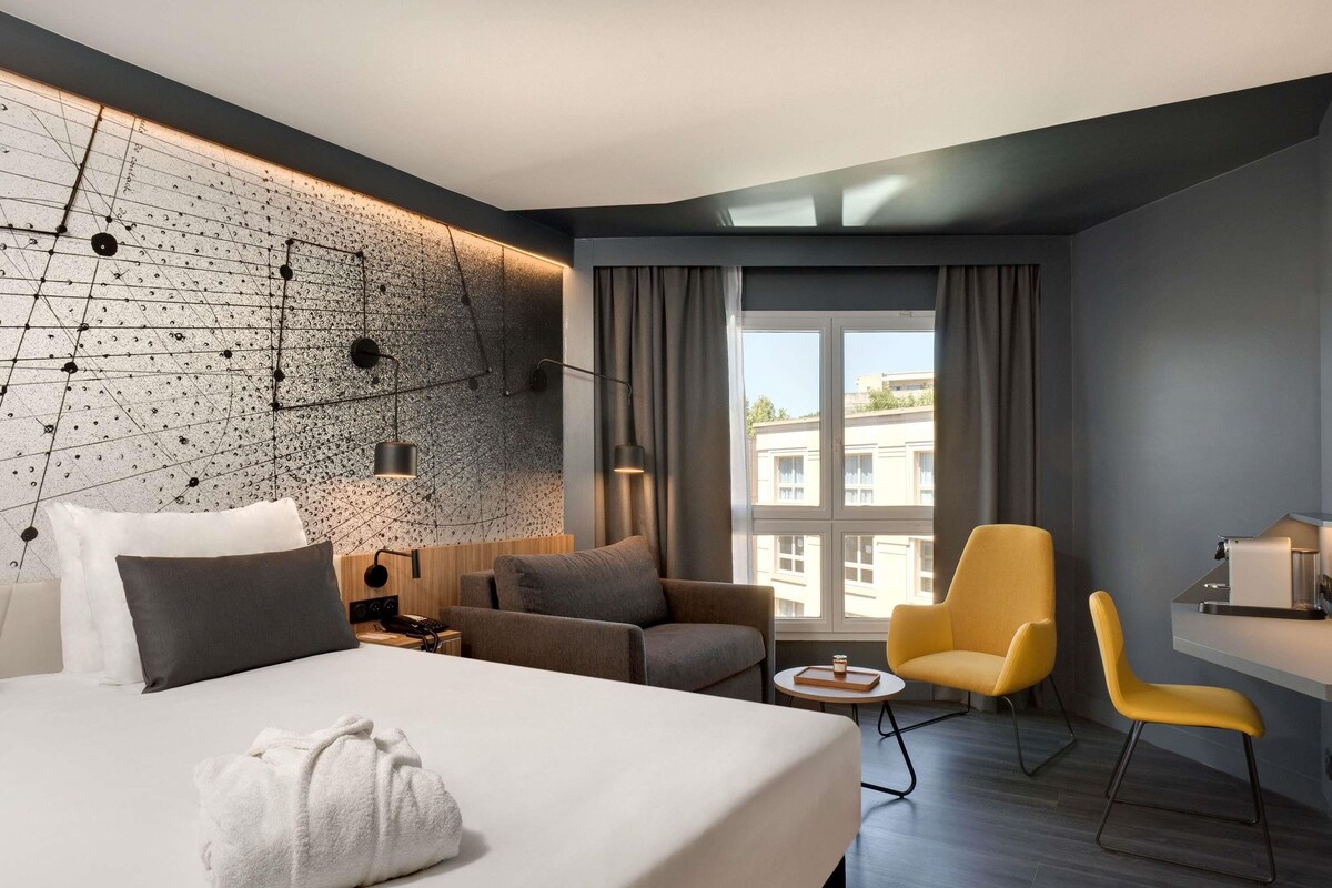 A modern hotel room with a large bed, a sofa, a desk, and a chair. There is a window with curtains, a wall with a geometric pattern, and a view of buildings outside.