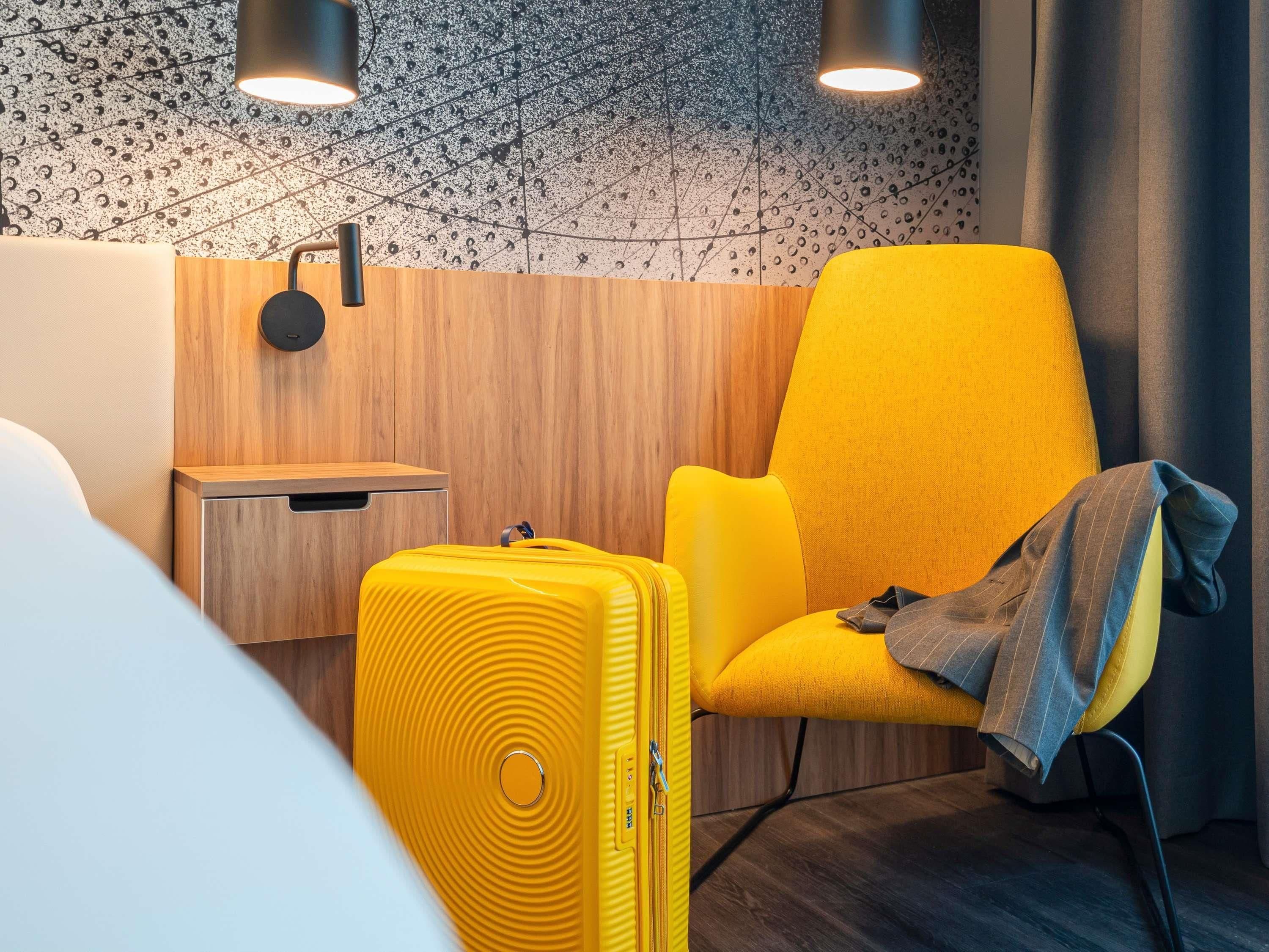 Habitación de hotel con pared de madera, un sillón amarillo, una mesita de noche pequeña y una maleta amarilla.