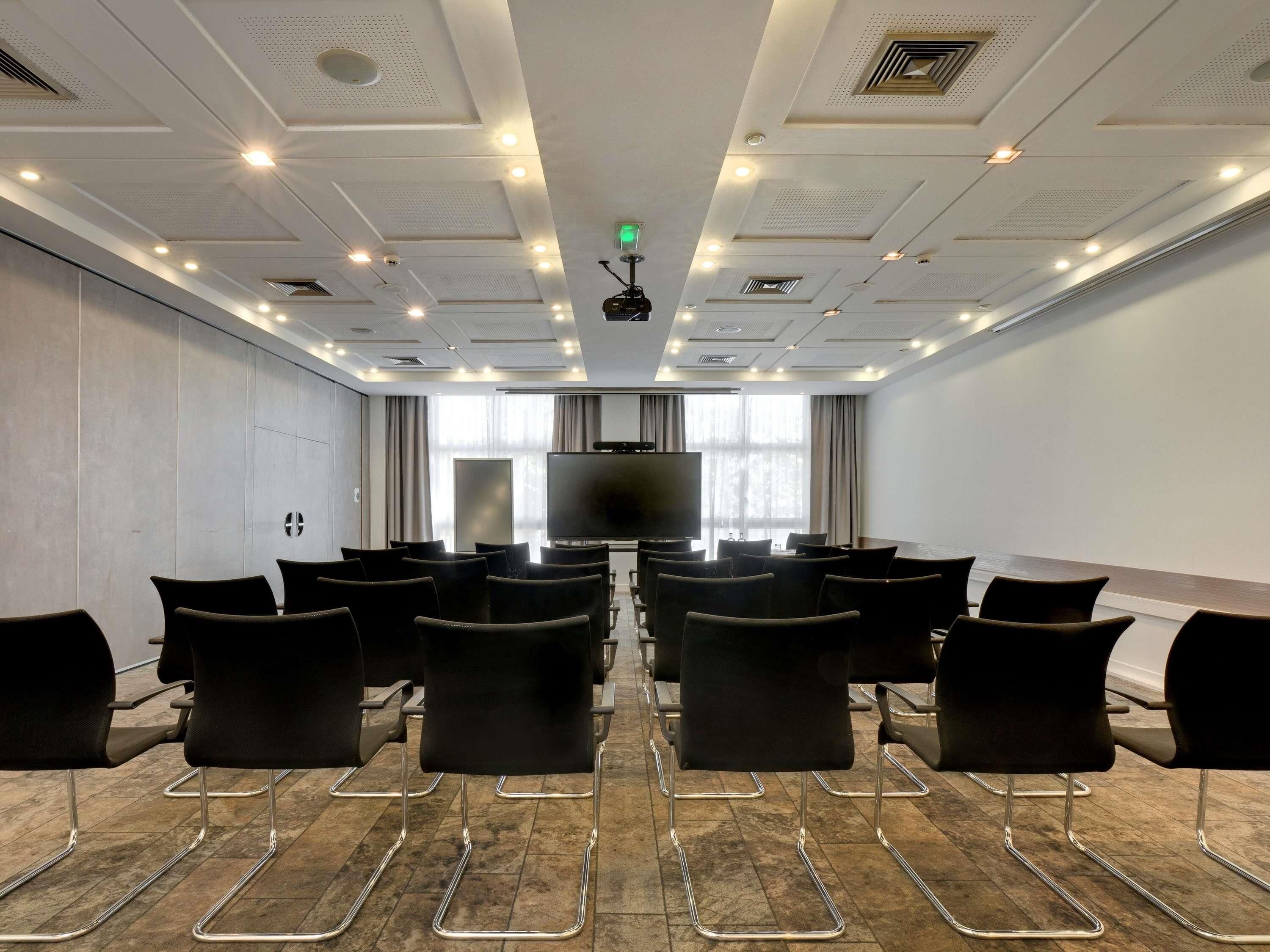 A conference room with a large flat-screen TV, rows of black chairs, and a ceiling with recessed lighting and ventilation.