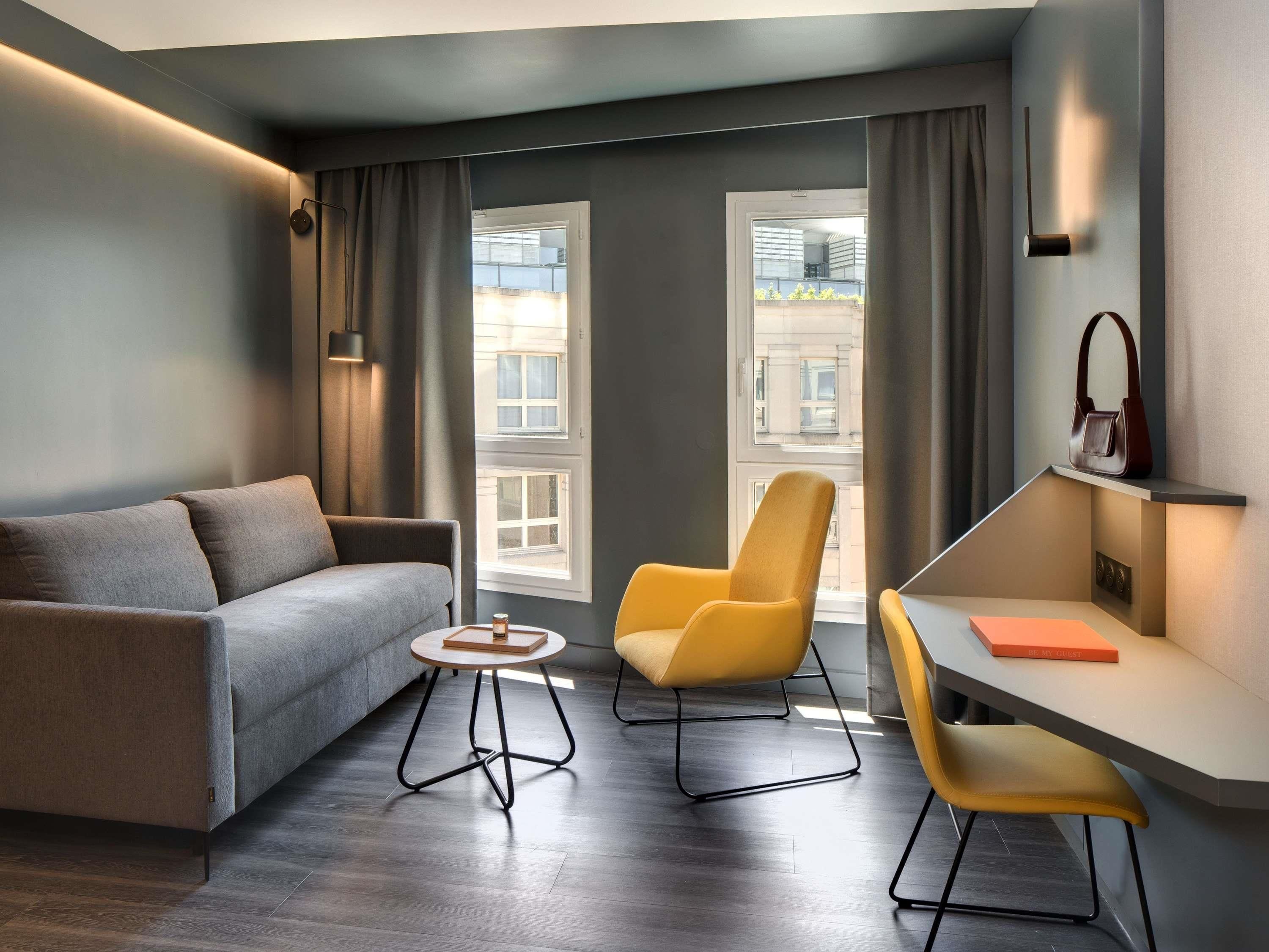 Una sala de estar moderna con un sofá, dos sillones, una mesita, un escritorio y vistas a la ciudad a través de amplios ventanales.