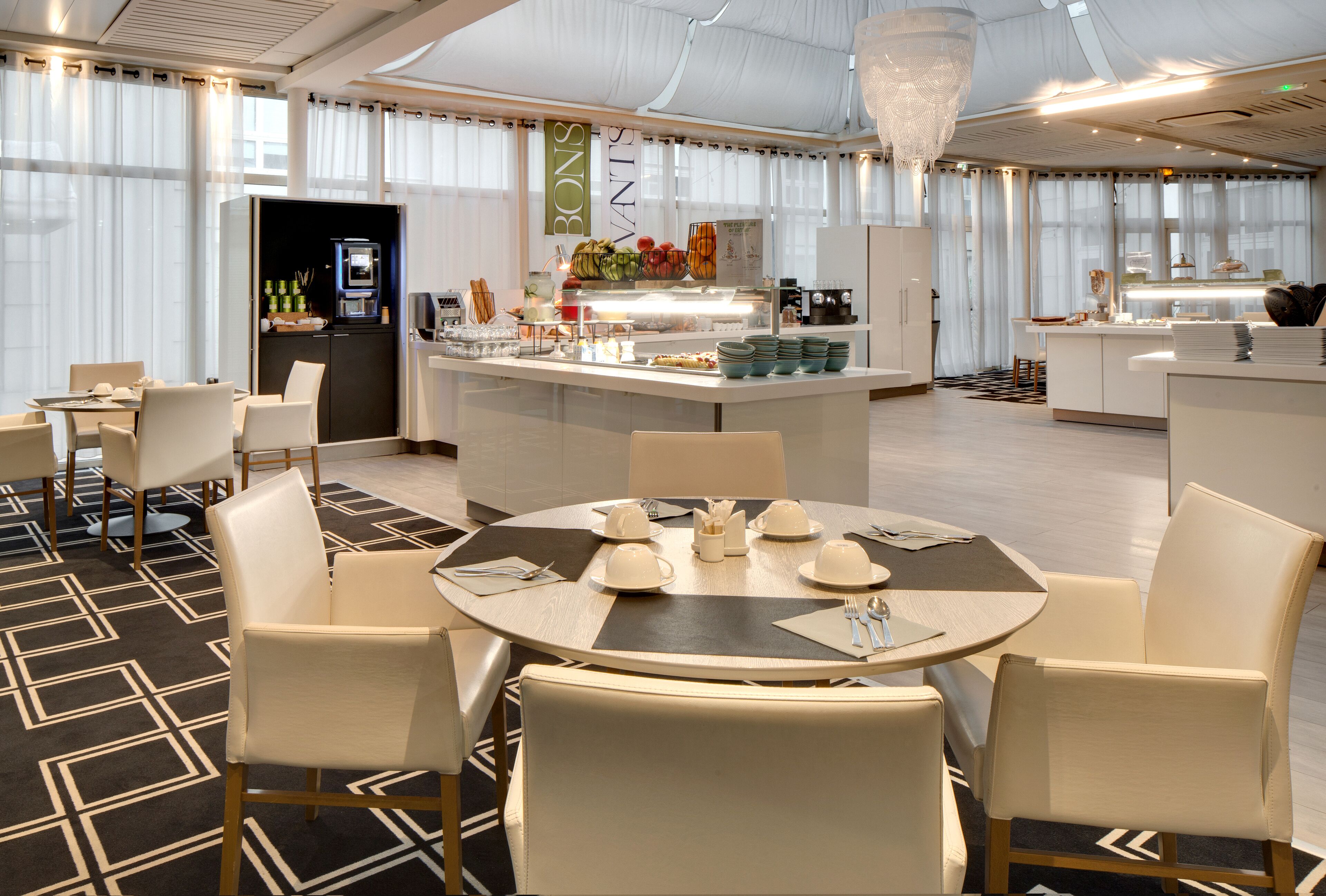Comedor de hotel con mesas redondas, sillas blancas, un mostrador de buffet y una araña.